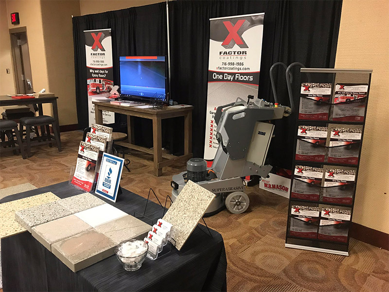 This image shows a trade show booth with flooring samples, marketing materials, a monitor, and a floor grinding machine. The banner reads "X Factor Coatings."