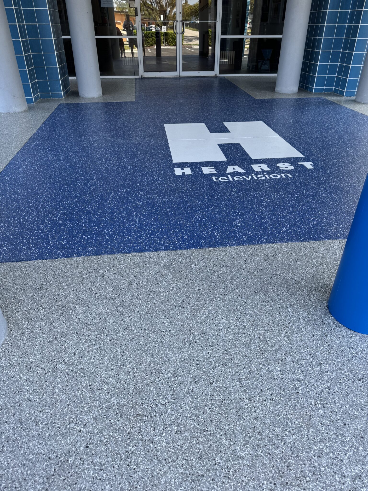 The image shows a blue-speckled floor with a large white "H" logo and "Hearst Television" text, in front of glass doors framed by blue tiles.