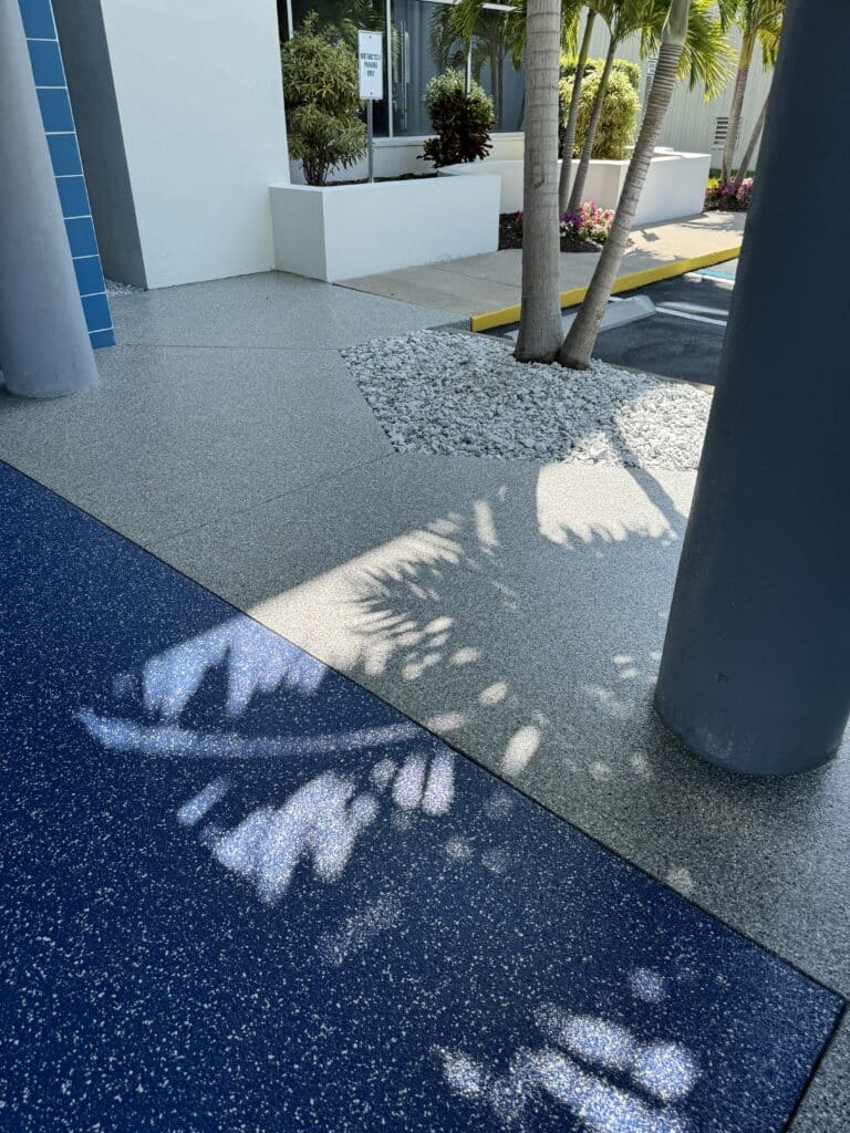 A modern building entrance with palm trees, casting shadow patterns on a textured walkway, accentuated by contrasting blue and grey ground surfaces.