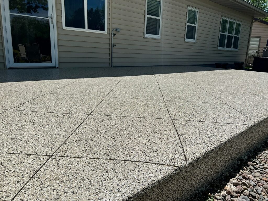 This image shows a large, textured concrete patio in front of a beige-sided house with windows and a sliding door, under a clear sky.