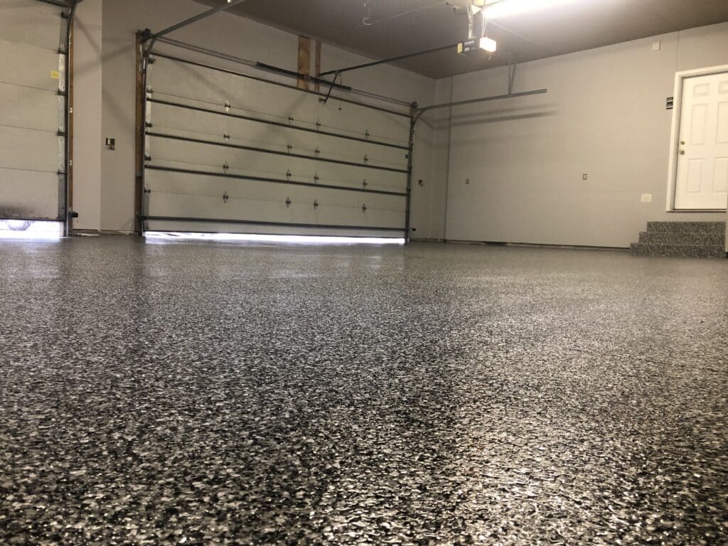 An empty garage with a closed door, speckled epoxy floor, and white walls. A small entry door and steps are visible on the right.