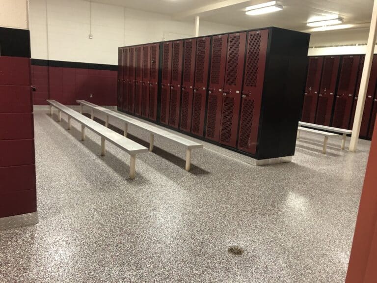 A locker room with red lockers, white benches, terrazzo flooring, and a black and white color scheme on the walls. It's well-lit and appears clean.