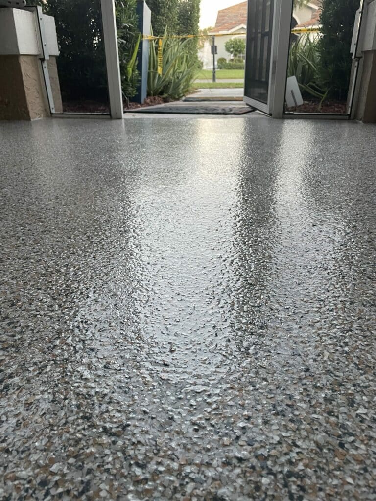 This image shows a speckled, wet garage floor leading to an open gate with a view of a residential area, greenery, and a partly cloudy sky.