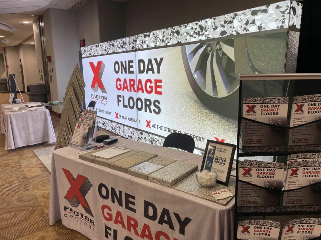 This image shows a promotional booth for "One Day Garage Floors" with a large banner, samples on the table, brochures, and award frames.