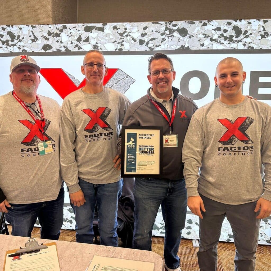 Four people stand smiling in front of a backdrop with a red "X" logo, wearing similar branded clothing and holding an accreditation plaque.
