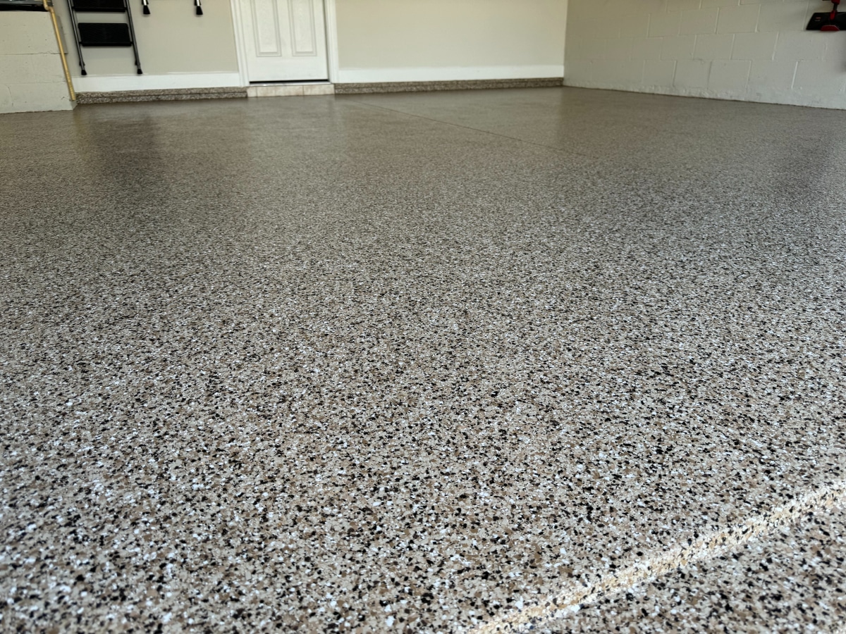 This image depicts a clean, speckled epoxy-coated garage floor with a smooth finish, leading to a closed white door and partial view of a wall.
