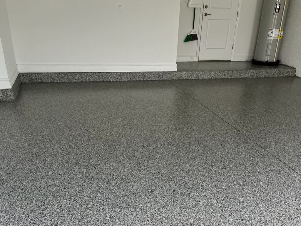 The image shows a room with gray speckled epoxy flooring, white walls, a baseboard, a door, a broom, and a water heater in the corner.