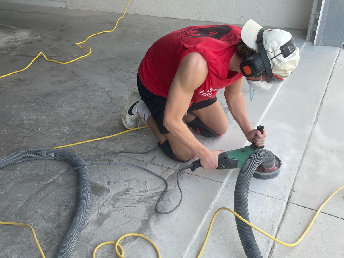 A person is kneeling on a concrete floor, using power tools for surface preparation. They wear a red shirt, hat, ear protection, and a face mask.