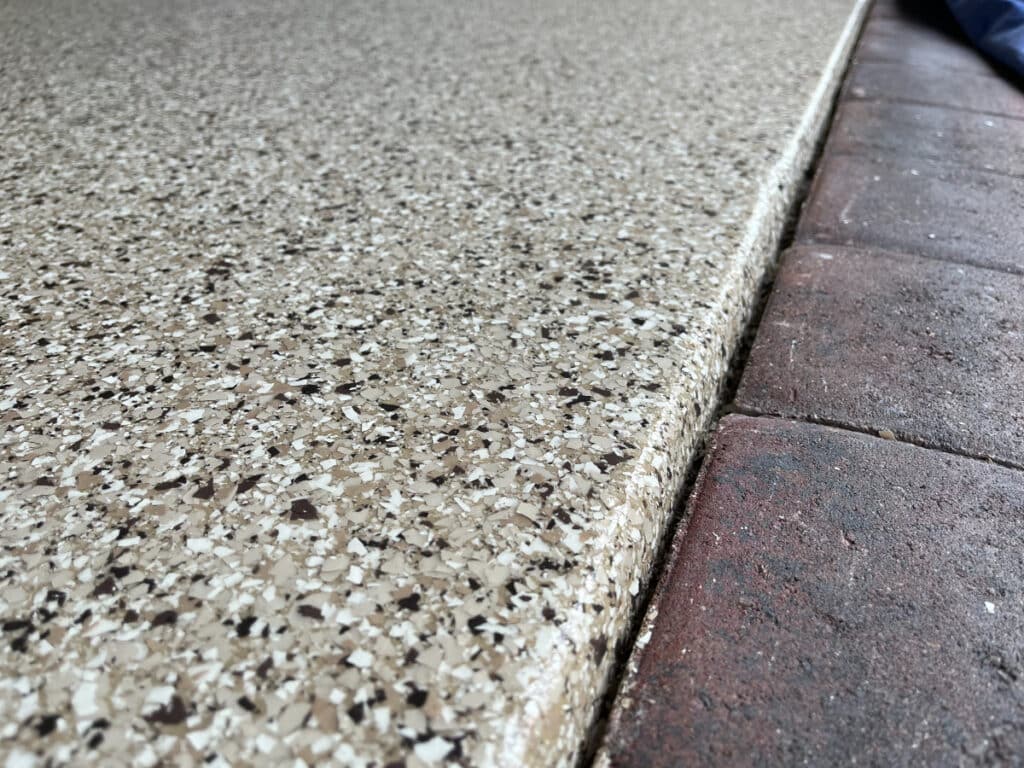 The image shows a close-up of a speckled floor meeting a brick edge, with a partial view of a person's leg to the right side.