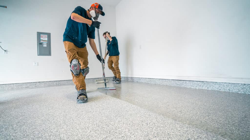 Two people in protective gear are applying a coating to a textured floor in a bright, empty room with white walls and a window.