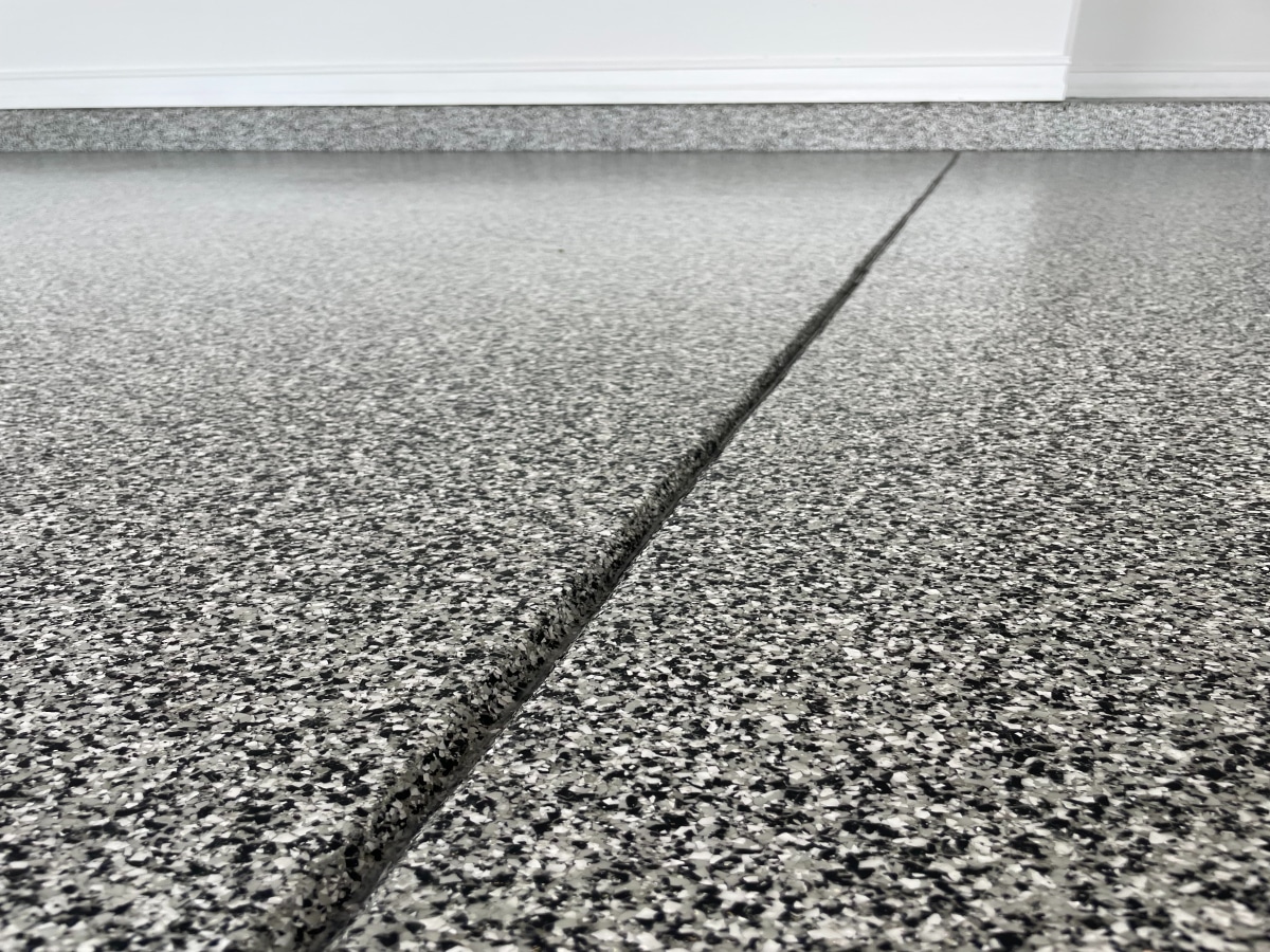This image shows a close-up view of a speckled gray epoxy floor with a visible control joint, juxtaposed against a white baseboard and wall.