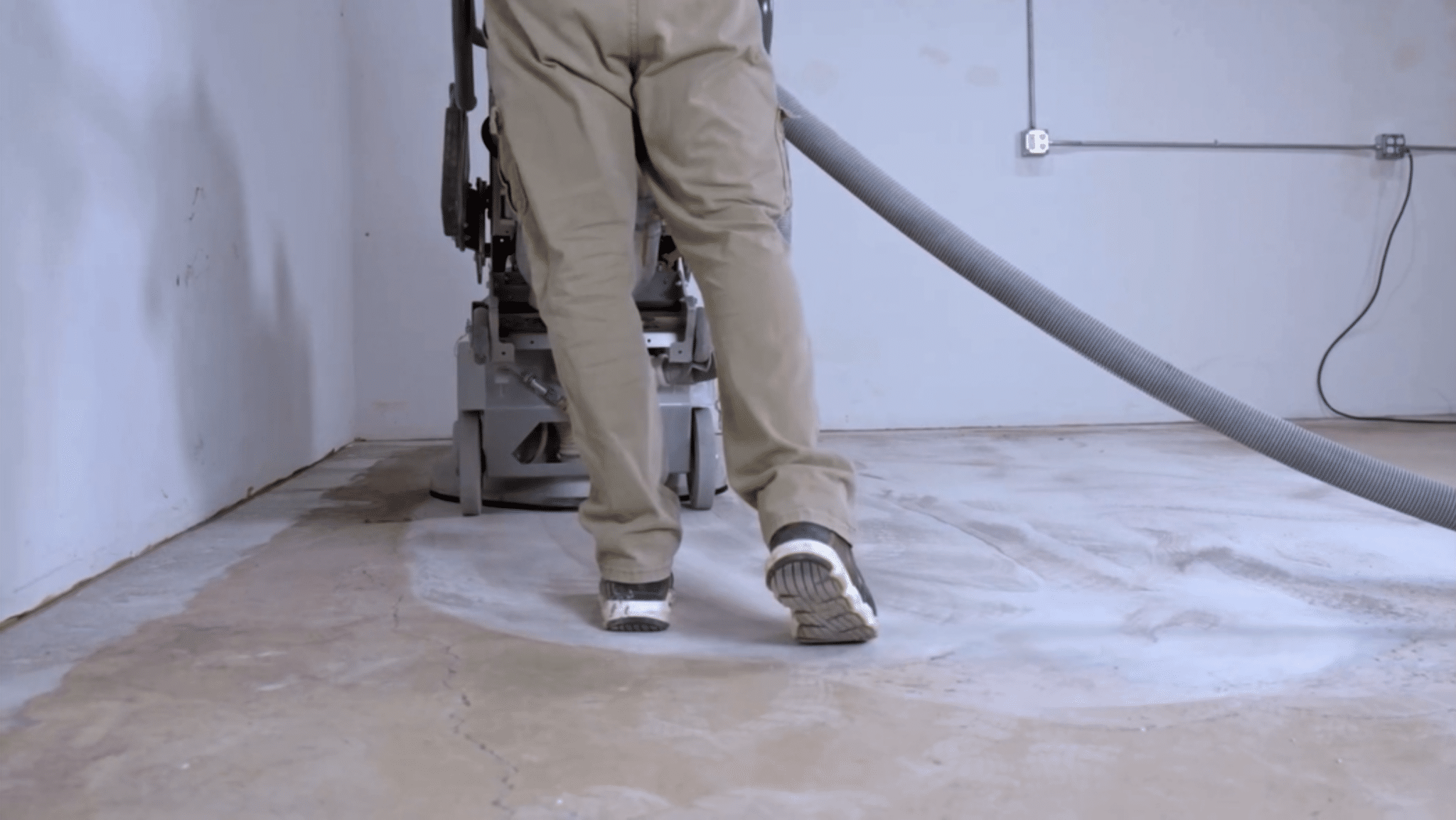 A person operates a large floor-cleaning machine with a hose in a room with a concrete floor, wearing tan work clothes and sneakers.