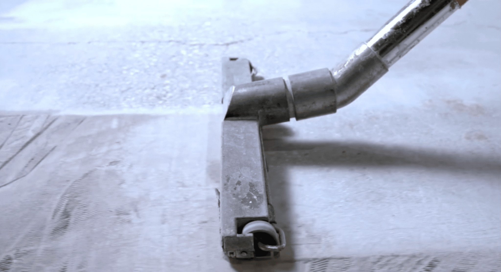 Metal vacuum cleaner nozzle on a textured floor, showing signs of wear and tear, focusing on its angled tube and flat head.