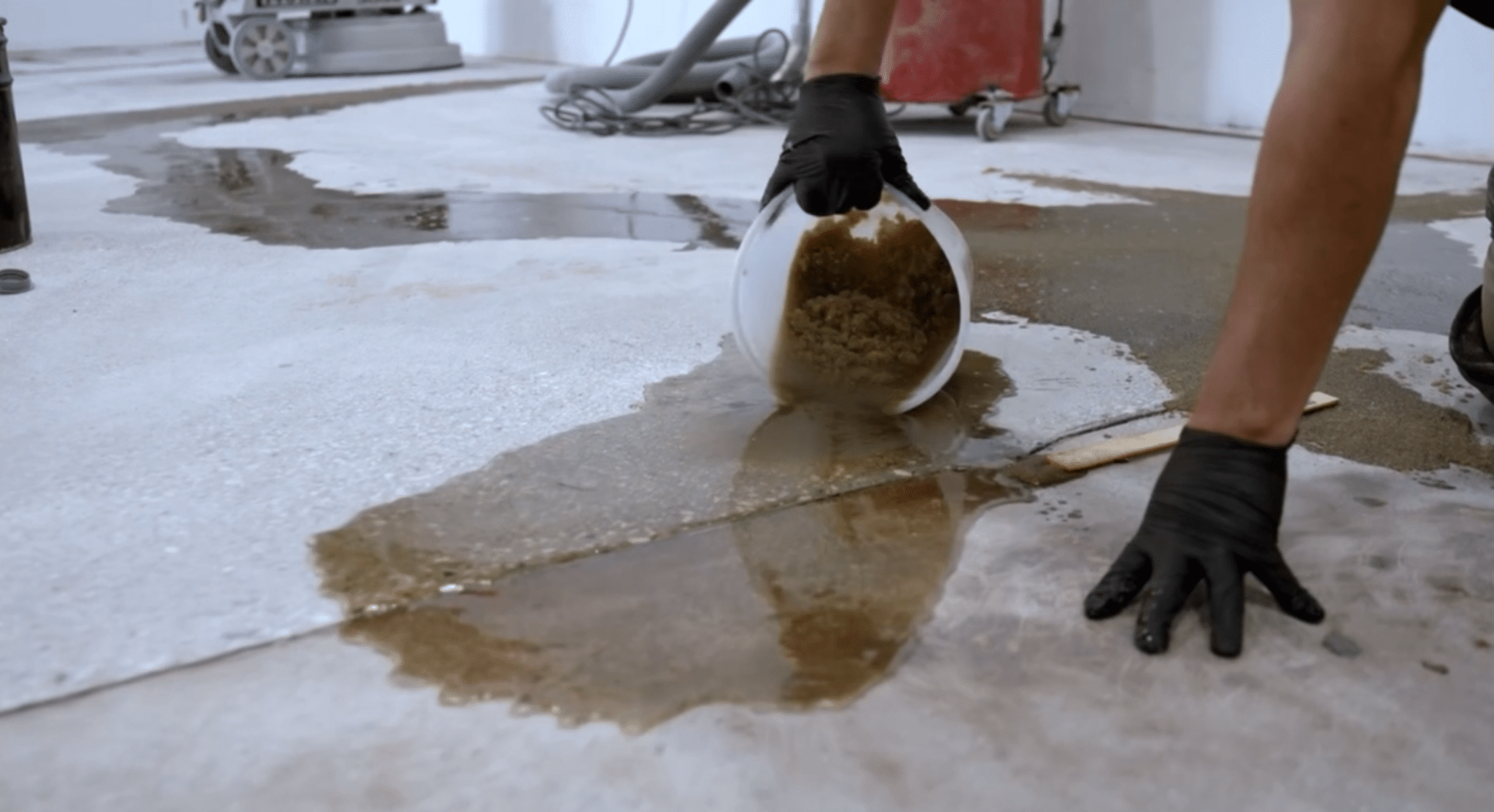 A person in gloves is pouring a substance onto a wet concrete floor, likely performing some type of construction or renovation work.