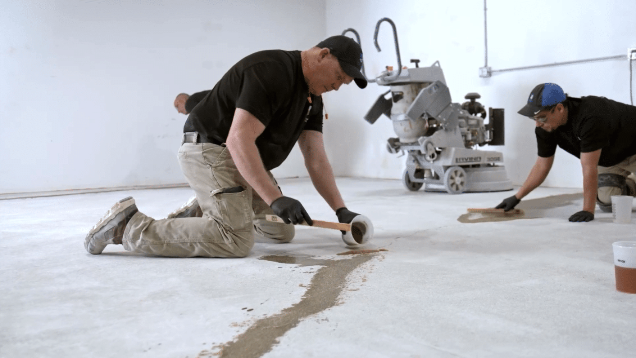 Two people are working on a concrete floor, one applying tape and another spreading a compound. Construction equipment is visible in the background.