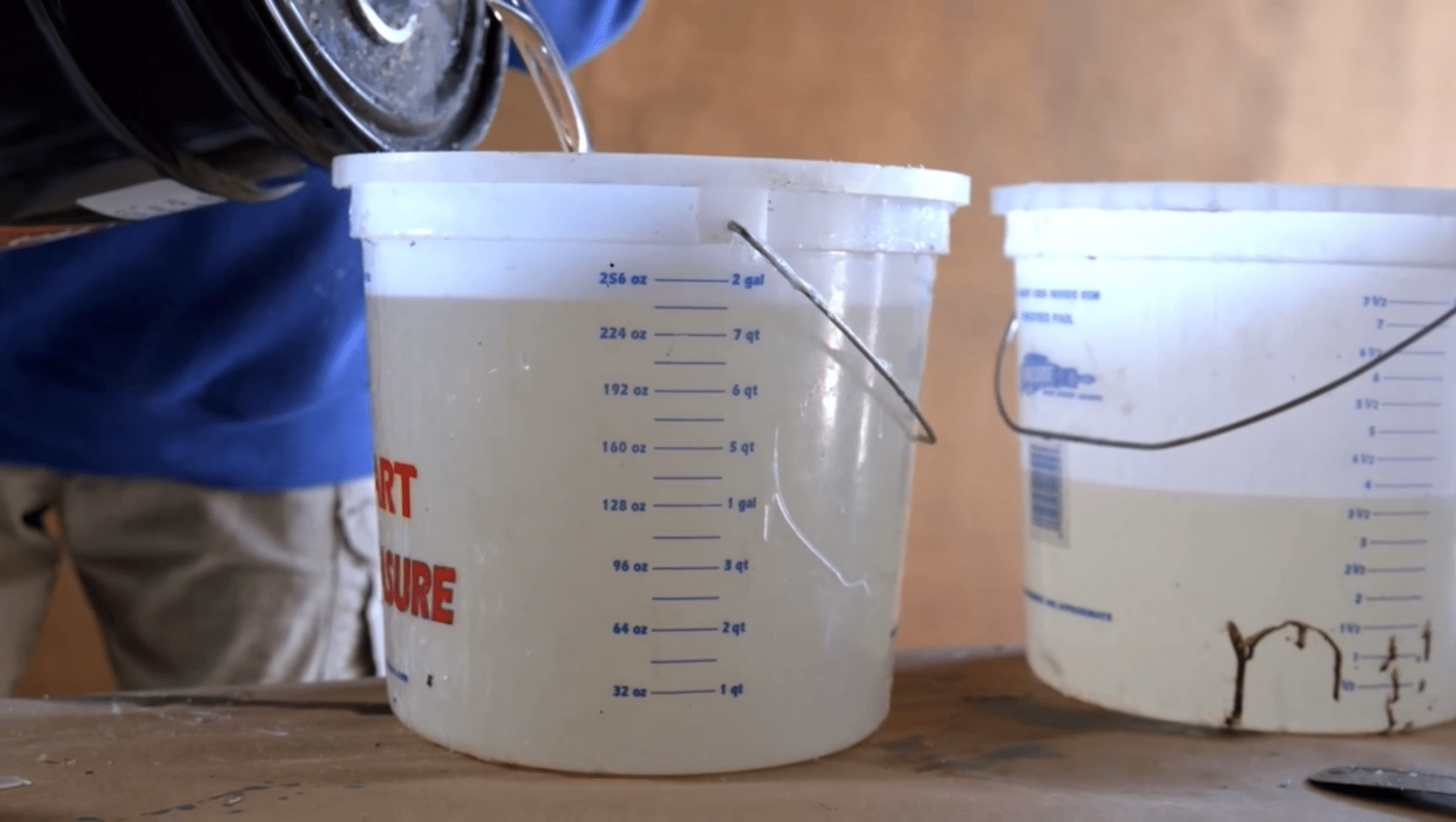 Two white graduated measuring buckets are on a surface; one is being filled with a liquid from a black container by a person.