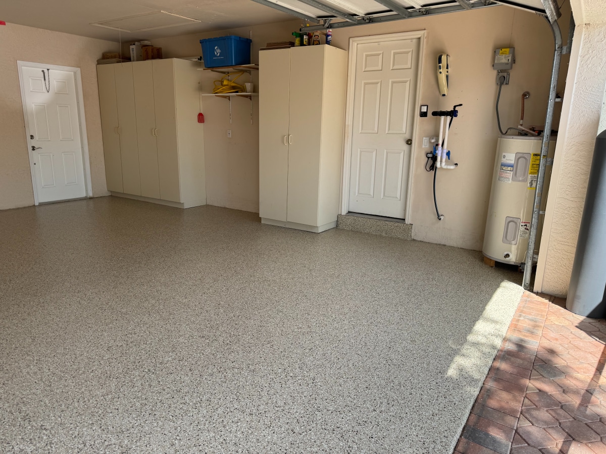A spacious, clean garage with a speckled floor, white cabinets, water heater, and a central vacuum system. Natural light enters through the open doorway.